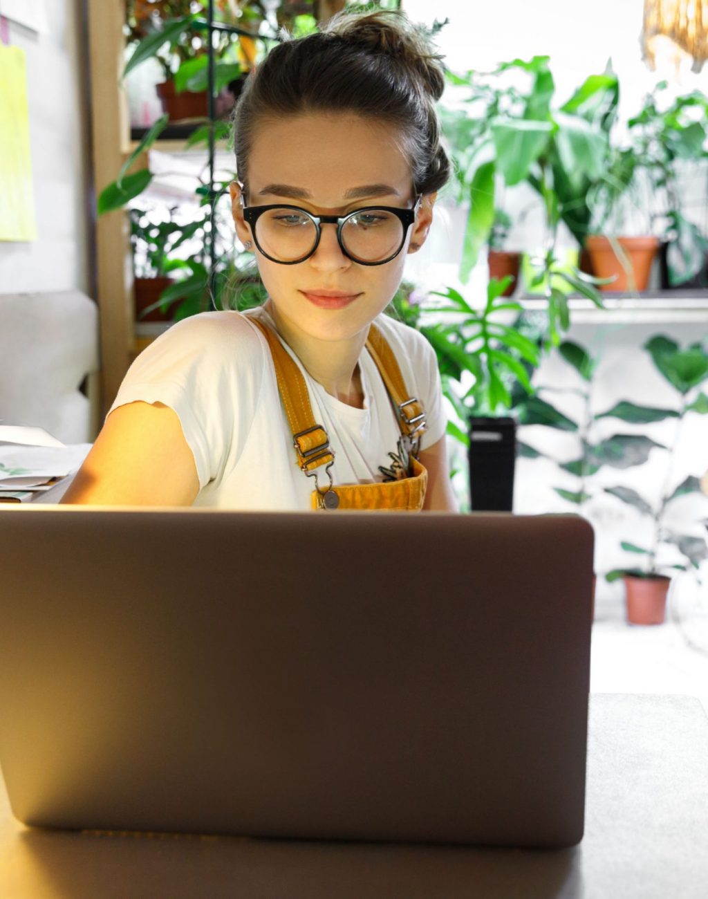 Young female gardener in glasses using laptop, communicates on internet with customer in home garden/greenhouse, reusable coffee/tea mug on table.Cozy office workplace, remote work, E learning concept